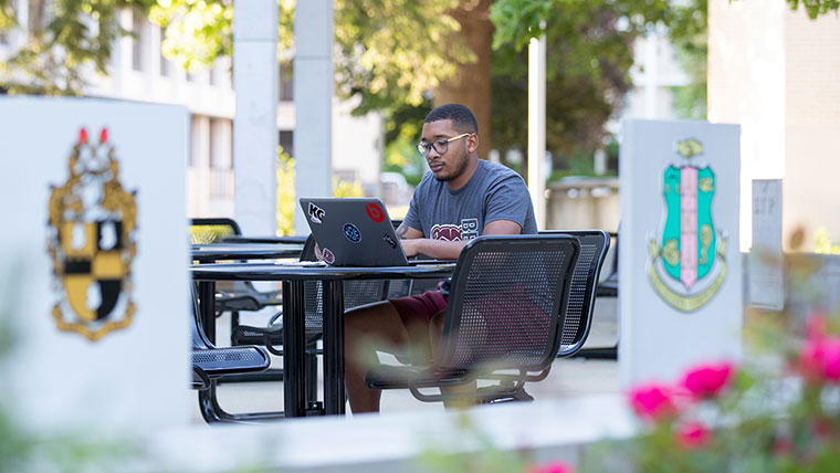 A student using his laptop while sitting in a campus courtyard.